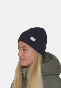 Schwarze Strickmütze mit angepasstem Design, die auf der Vorderseite ein kleines weißes Etikett aufweist. Getragen zu einer mittel langen blonden Frisur.