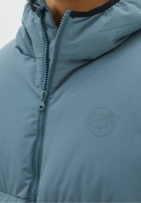 Ljusblå vadderad jacka med dragkedja, utrustad med huva och en diskret logotypdetalj på bröstet. Slät yta med matt finish.