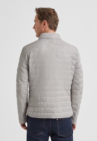 Hellgraue Steppjacke mit Kragen, ausgestattet mit horizontalen Steppnähten und einem Etikett am Rücken. Das Kleidungsstück hat lange Ärmel und eine taillierte Passform.