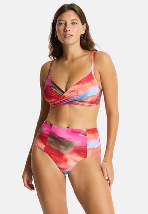 Kobieta stoi przodem, ubrana w bikini z wysokim stanem o kolorowym, abstrakcyjnym czerwono-różowo-niebiesko-brązowym wzorze na jednolitym tle.