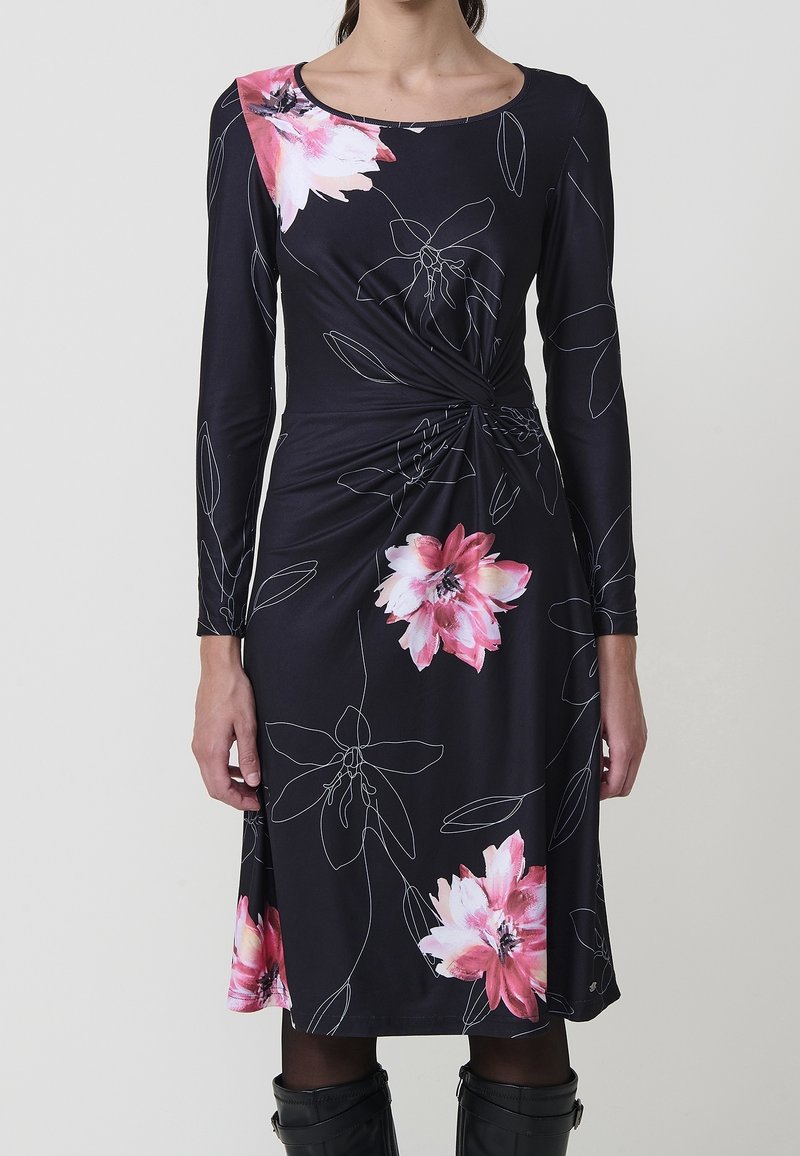 Femme portant une robe noire à manches longues avec des imprimés floraux roses et blancs, détail froncé à la taille, associée à des collants noirs et des bottes.