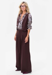 Brązowe szerokie spodnie w prążki w połączeniu z bluzką w kwiatowy wzór i brązową kamizelką, do tego duże okulary i eleganckie buty.