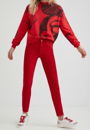 Osoba ubrana w czerwone, obcisłe jeansy z wysokim stanem, czerwoną bluzę z czarnymi abstrakcyjnymi wzorami oraz białe trampki z czerwonymi sznurowadłami.