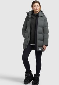 Cappotto grigio imbottito con cappuccio, chiusura a zip e polsini elasticizzati. Indossato sopra un maglione nero e leggings, abbinato a stivali neri con lacci.