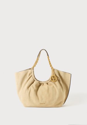 Bolso beige de ante con parte superior fruncida, asas de cadena dorada y charm redondo dorado con el logo MK sobre un fondo liso.