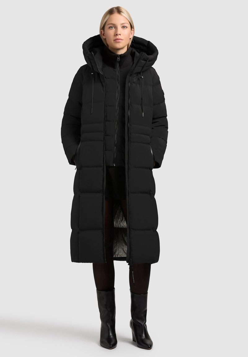 Sieviete, tērpusies garā melnā pūkainā mētelī ar kapuci, melnā apvalka iekšējā slānī ar rāvējslēdzēju, melnās zeķbikses un melnās potītes zābakos, stāv uz pelēka fona.