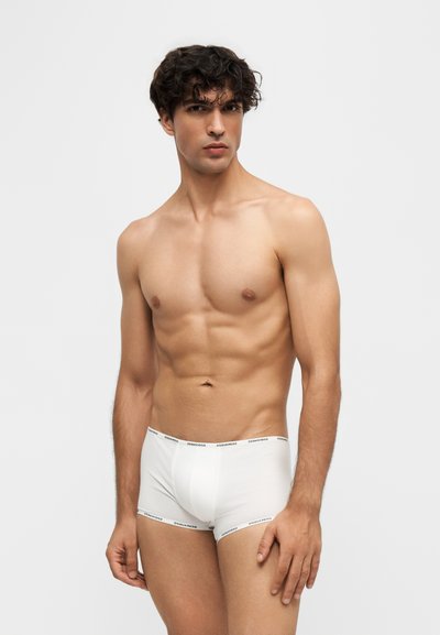 Jeune homme aux cheveux bouclés foncés, portant un boxer blanc, debout devant un fond uni gris clair.