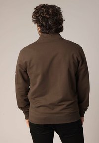 Sweatshirt marron en tissu doux, avec un col montant et des poignets côtelés, et une poche sur la manche gauche.