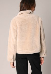 Chaqueta de forro polar beige, corta, con una textura suave y un amplio cuello. Presenta un dobladillo recto y costuras terminadas, combinada con pantalones negros.