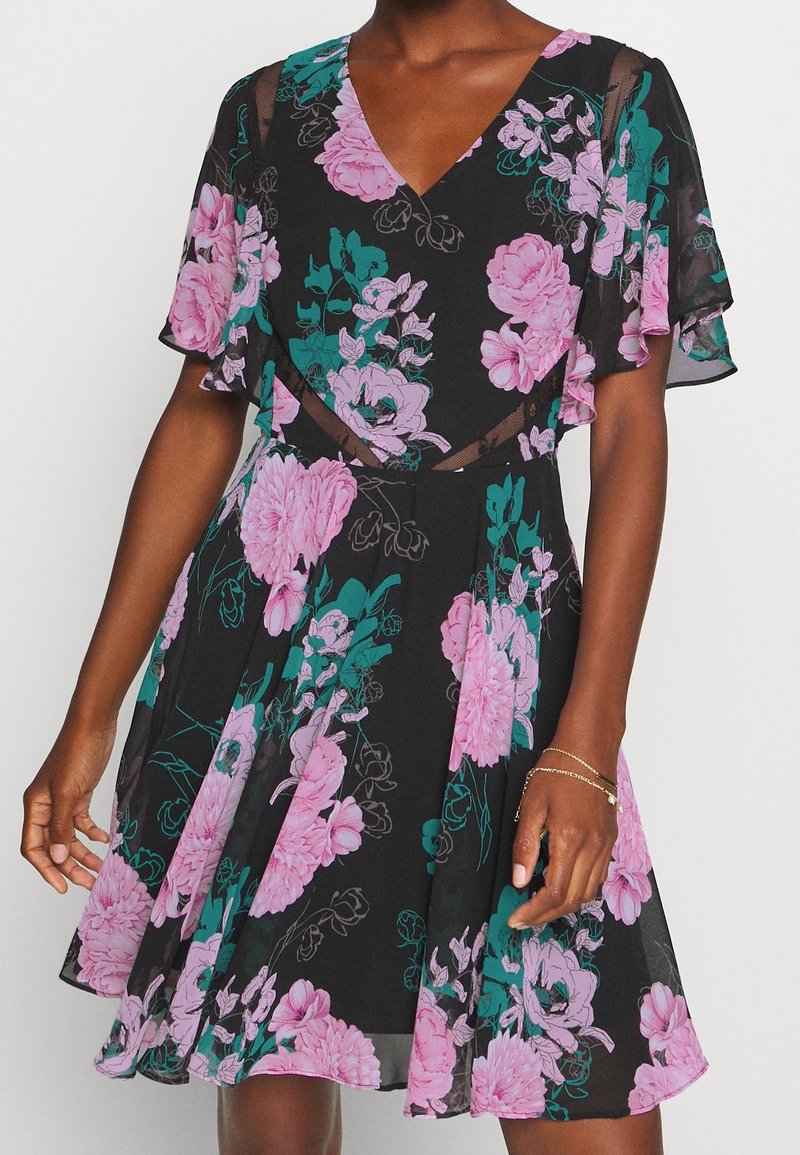 Femme portant une robe noire avec un motif floral rose et vert et des manches volantées, debout devant un fond blanc.