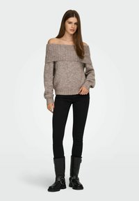 Pull en tricot à épaules nues de couleur marron clair avec un col en côtes ; associé à un jean skinny noir et des bottes noires hautes avec des accents métalliques.