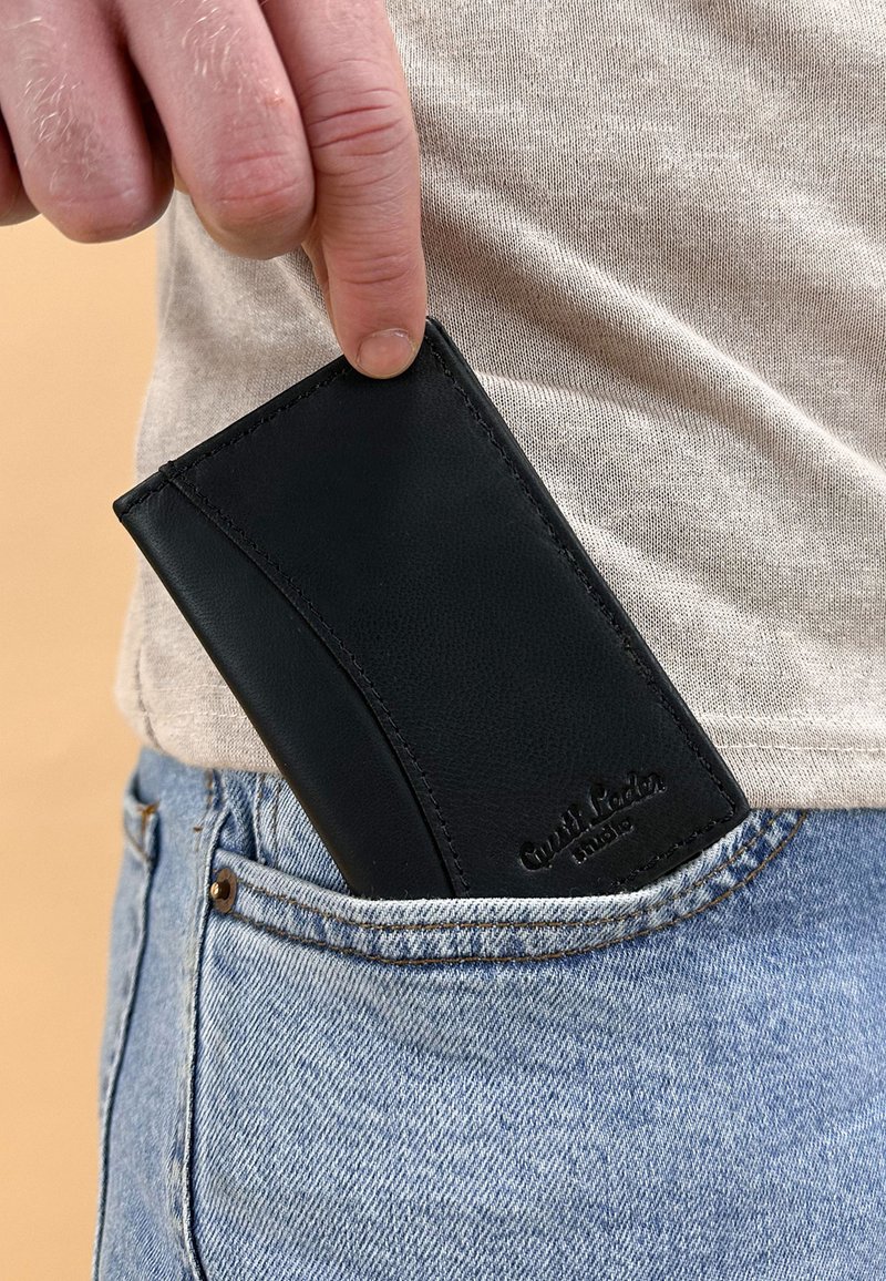 Hand steckt eine schwarze Lederbrieftasche in die Vordertasche einer blauen Jeans vor einem beigen Hemd und hellem Hintergrund.