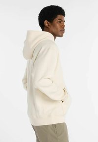 Hombre vistiendo sudadera con capucha de color crema, con las manos en los bolsillos, de pie de lado contra un fondo claro y uniforme.