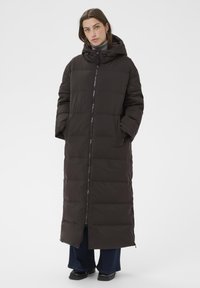 Pitkä puffer-takki tummanruskeana, jossa on huppu, vetoketju, tikattu pinta, sivutaskut ja suora siluetti.