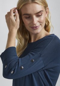 Blaues Langarmoberteil mit gerippten Bündchen, mit silbernen Knopfdetails an den Ärmeln, aus einem weichen, strukturierten Stoff.