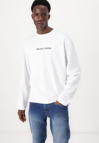 Hvit langermet t-skjorte med "RED BULL RACING" trykt i svart over brystet, sammen med blå jeans.