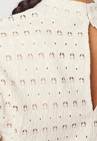 White textured fabric featuring circular eyelet patterns and small holes, with a keyhole back design. Soft and lightweight material.