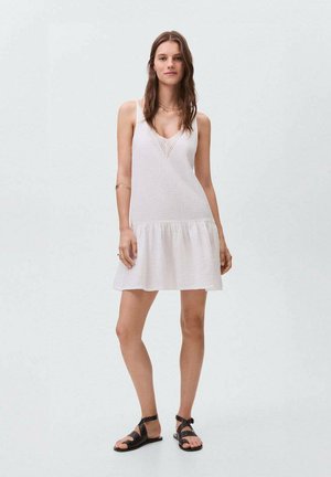 Robe d'été blanche légère avec un tissu texturé, décolleté en V et jupe évasée. Associée à des sandales noires à lanières.