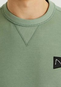 Primo piano di una persona che indossa una felpa a girocollo verde chiaro con dettagli a V a coste e una piccola etichetta nera sul petto.