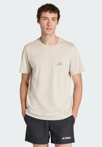 Jasnobeżowa koszulka z okrągłym dekoltem i krótkimi rękawami, z małym szarym logo Adidas na lewej piersi. Noszona z czarnymi szortami.