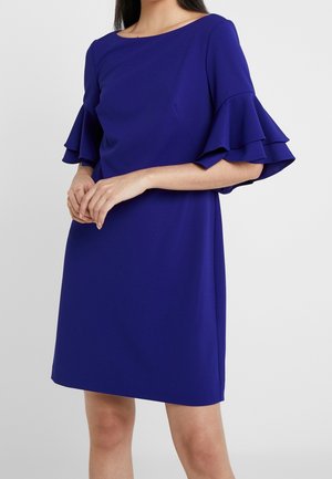 Nainen, jolla on polvenmittainen koboltinsininen mekko, jossa on kerrokselliset levenevät lyhyet hihat ja leveä pääntie, seisoo tasaisen taustan edessä.