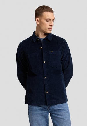 Jeune homme aux cheveux courts portant une chemise en velours côtelé bleu foncé boutonnée et un jean bleu clair, regardant vers le bas avec les mains derrière le dos.