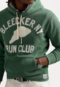 Grön hoodie med "BLEECKER NY RUN CLUB" i vit text. Har en framlapp, dragsko i huven och strukturerad tyg. Vit logotypaccent.