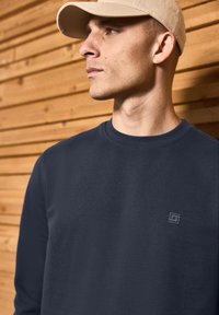 Navy blue crew neck sweatshirt made of cotton with a subtle logo on the chest. The fabric appears smooth against a wooden background.