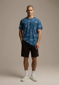 Niebieska koszula z krótkim rękawem w kwiatowy wzór i kieszenią, zestawiona z ciemnymi szortami i białymi sneakersami. Tło jest neutralne i gładkie.