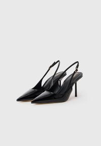 Un paio di décolleté nere in vernice con punta affusolata e cinturino alla caviglia, accompagnate da sottili tacchi a stiletto su sfondo bianco.