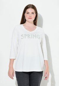 Camiseta blanca de manga larga con escote en V que presenta "SPRING" escrito en rhinestones verdes. Tejido suave con un ajuste relajado, combinado con pantalones gris oscuro.