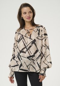 Losse beige blouse met een zwart abstract patroon, voorzien van een V-hals met strikdetail en elastische manchet. Glad stof met een subtiele glans.