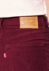 Burgundyröda corduroybyxor med en strukturerad yta, utrustade med en läderpatch i tan och en liten röd logotypetikett på bakfickan.