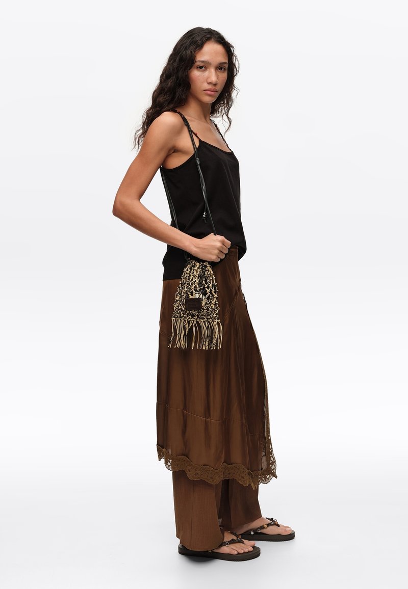 Mujer con cabello largo y rizado, que lleva una camiseta negra sin mangas, falda marrón sobre pantalones, sandalias y sostiene una bolsa de hombro tejida con flecos.