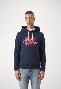 Felpa blu navy in misto cotone con tasca a marsupio frontale, coulisse e una grafica rossa con testo bianco. Forma casual, polsini a costine.