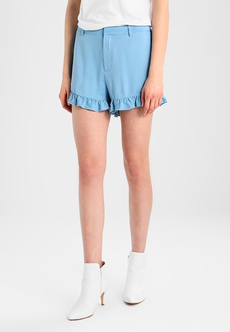 Shorts bleu clair avec un ourlet volants, fabriqués dans un tissu doux. Présente un devant plat et des poches latérales. Associés avec des bottines blanches.