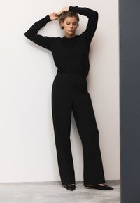 Pull noir à manches longues associé à un pantalon noir taille haute à jambes larges. Le mannequin se tient contre un mur clair, portant des chaussures noires à bout pointu.