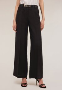 Wijdvallende zwart pantalon van gladde stof, met een hoge taille, plooien en een smalle zwarte riem met een zilveren gesp.