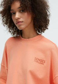 Peach sweatshirt with a crew neck, oversized fit, and "SENSES . THE LABEL" printed in a lighter shade. Soft texture, casual design.