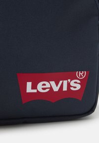 Borsa in tessuto blu navy con un logo Levi's rosso ben visibile, con testo bianco, evidenziato da una forma curva alla base del logo. Superficie testurizzata.