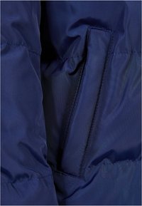 Giacca a vento blu navy in tessuto con una texture liscia, dotata di una tasca laterale visibile e cuciture rinforzate lungo le giunture.