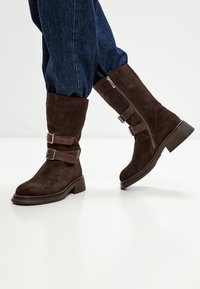 Botas de tobillo de ante marrón con dos hebillas decorativas, puntera redondeada y un bajo tacón bloque. Cuenta con una cremallera lateral para facilitar su uso.
