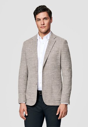 Mężczyzna ubrany w beżową, teksturowaną marynarkę na białej koszuli zapinanej na guziki i ciemnych spodniach, stojący na tle jasnego, gładkiego tła.