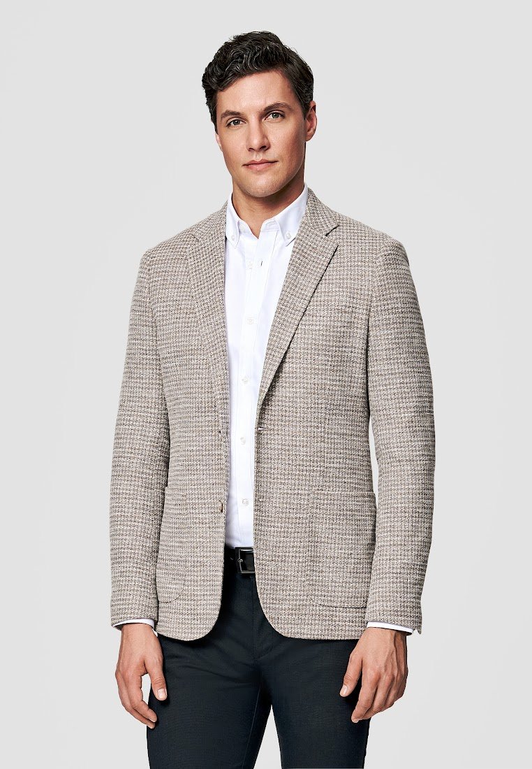 Mężczyzna ubrany w beżową, teksturowaną marynarkę na białej koszuli zapinanej na guziki i ciemnych spodniach, stojący na tle jasnego, gładkiego tła.