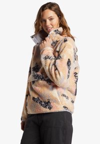 Fleece-pullover i pastelfarver med sorte abstrakte mønstre, høj krave og lynlåslukning; blød, tekstureret overflade.