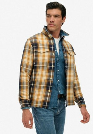Camicia in flanella a quadri nei toni del giallo, blu navy e bianco; presenta due tasche sul petto, chiusura con bottoni e una camicia di jeans a strati sotto.
