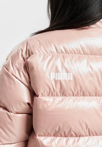 PUMA pūkinis švarkas šviesiai rožinės spalvos, su blizgiu paviršiumi, siuvinėjimais ir balta logotipo etikete ant nugaros, pabrėžiančiu jo elegantišką dizainą.