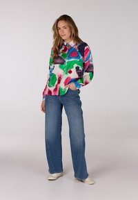 Kleurrijke blouse met abstracte patronen in roze, groen en blauw, gecombineerd met wijd uitlopende blauwe jeans en witte sneakers.