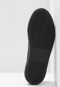 Svart sneaker med en texturerad gummisula med ett sicksackmönster, övre i tyg och en låg profil, lutad mot en vit bakgrund.