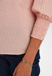 Blusa con motivo floreale in rosa chiaro con accenti rossi. Le maniche a sbuffo presentano una texture arricciata. Il modello indossa un braccialetto a catena e jeans scuri.